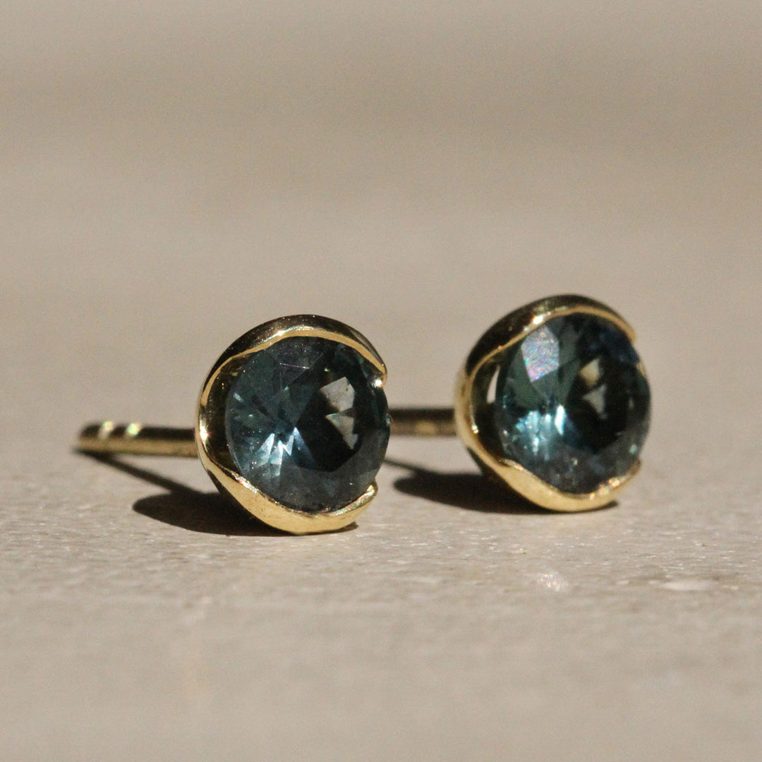 Sophie Hughes Poppy Bezel Montana Sapphire Studs in 18k Yellow Gold featuring a pair of 4.75mm Dark Teal Round Montana Sapphires, from a slight angle