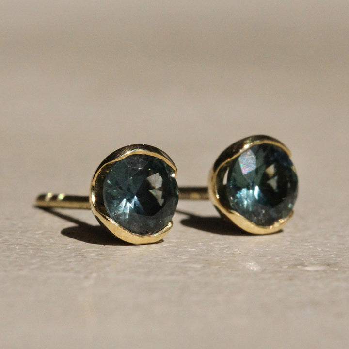 Sophie Hughes Poppy Bezel Montana Sapphire Studs in 18k Yellow Gold featuring a pair of 4.75mm Dark Teal Round Montana Sapphires, from a slight angle