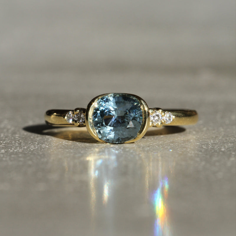 Sophie Hughes Poppy Bezel Ring in 18K Yellow Gold featuring a 1.5-ct oval cut light denim blue Montana Sapphire with 4 white melee diamonds
