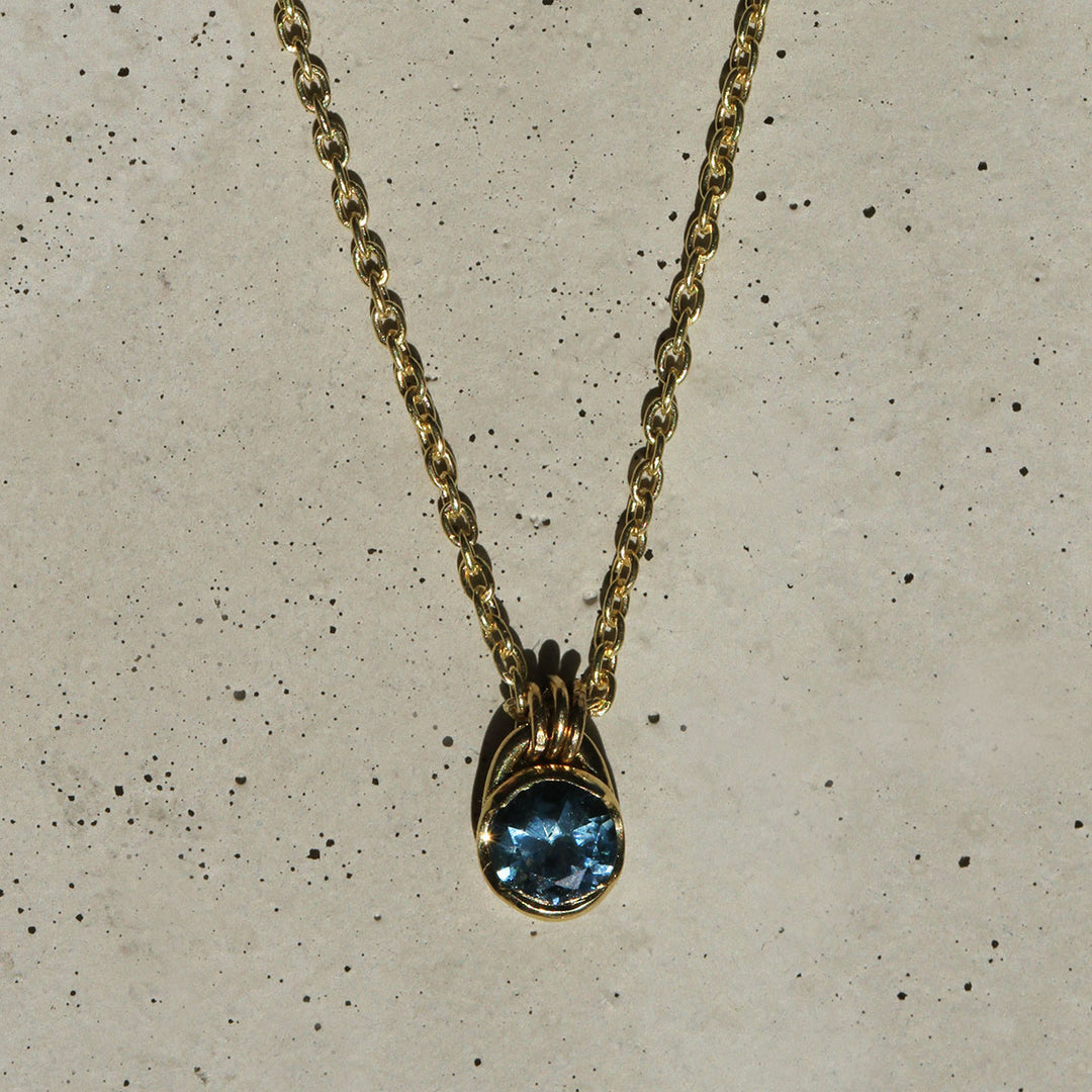 A blue Montana Sapphire necklace set in a yellow gold bezel, with the pendant hanging from a gold chain.