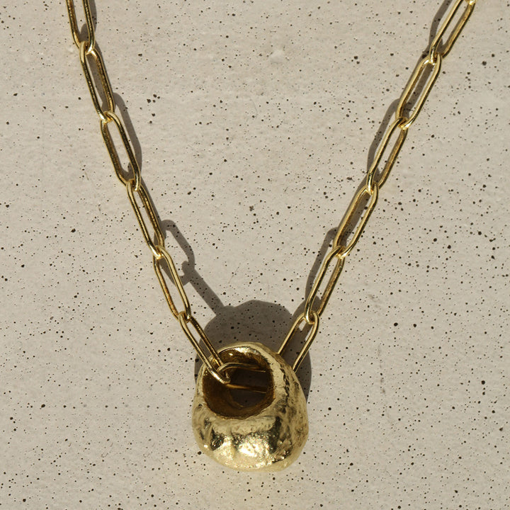 A solid gold 18k bell pendant swinging on a gold heavyweight Oblong link chain against a concrete background.
