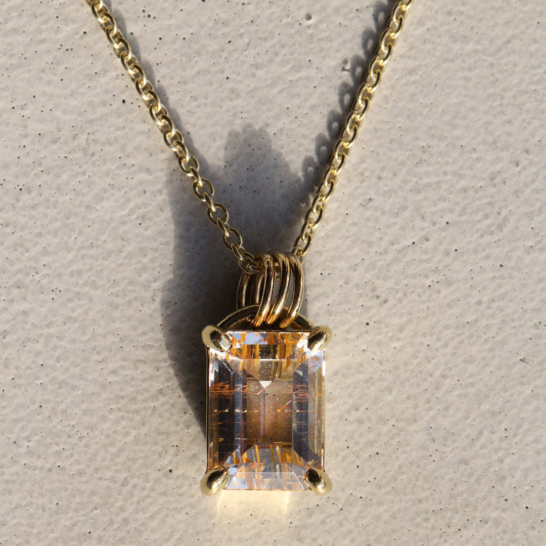 A rutilated quartz arch necklace with a colorless stone and natural golden rutile, suspended from a 18k yellow gold Arch Bail on a gold chain, displayed against a concrete background.