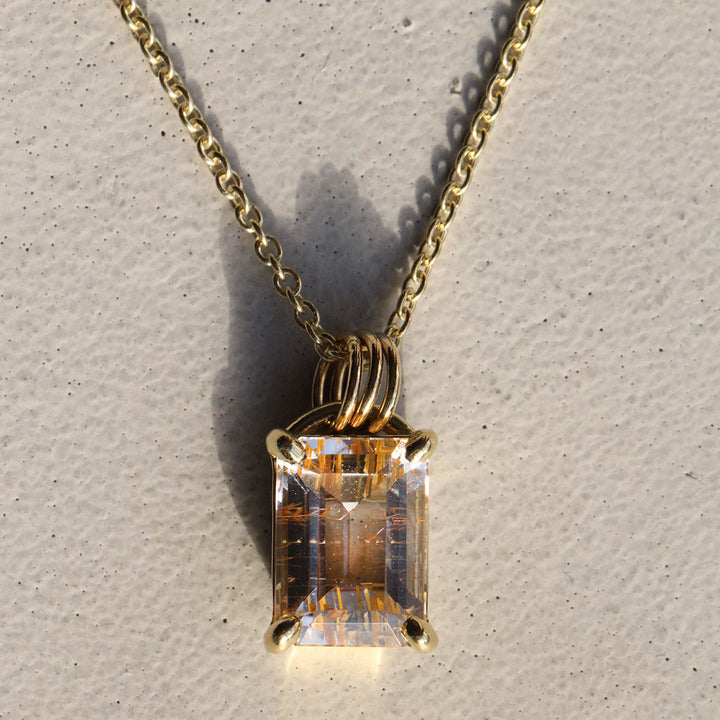 A rutilated quartz arch necklace with a colorless stone and natural golden rutile, suspended from a 18k yellow gold Arch Bail on a gold chain, displayed against a concrete background.