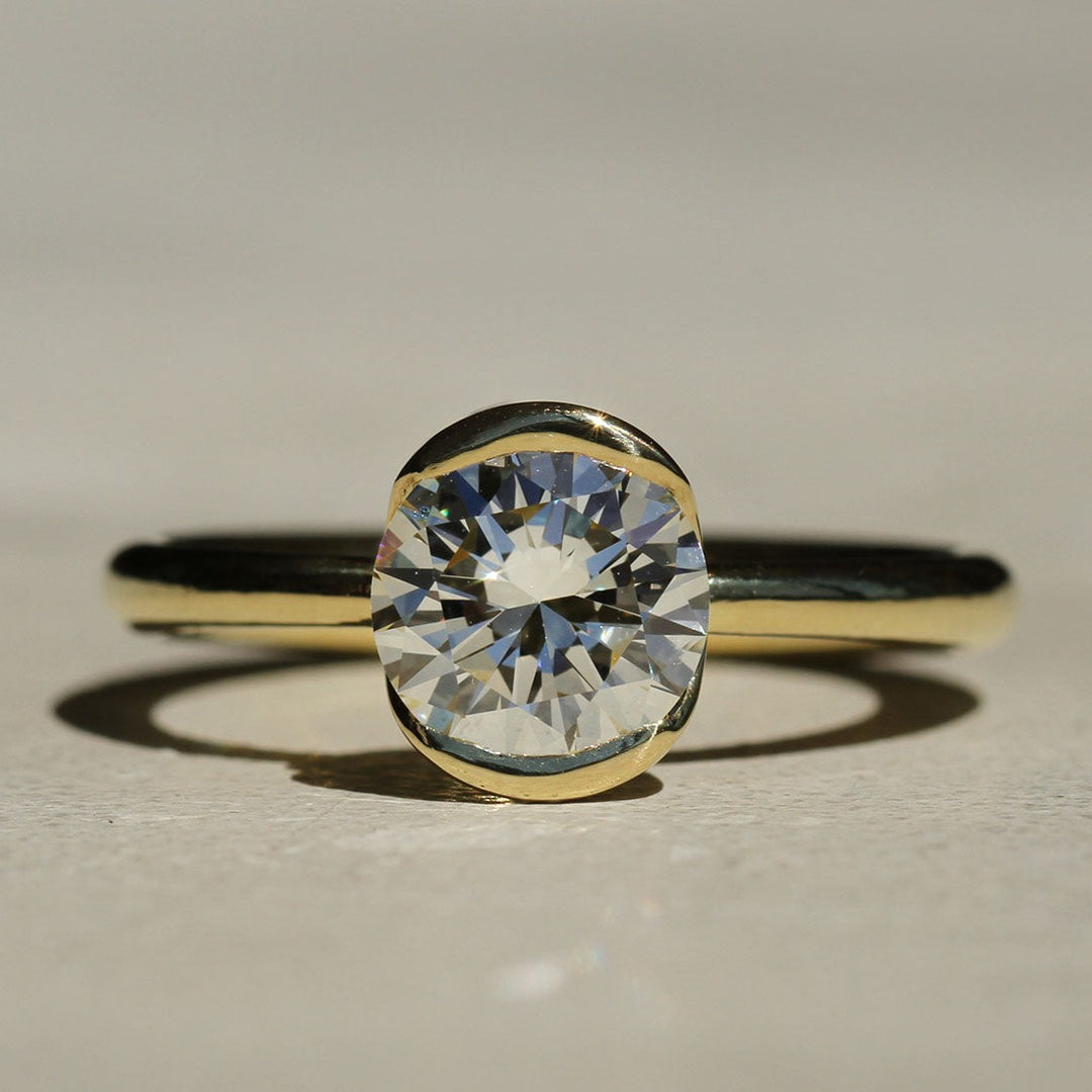 A close-up image of a diamond solitaire ring with a gold band, showcasing the round brilliant cut diamond set in a bezel setting.