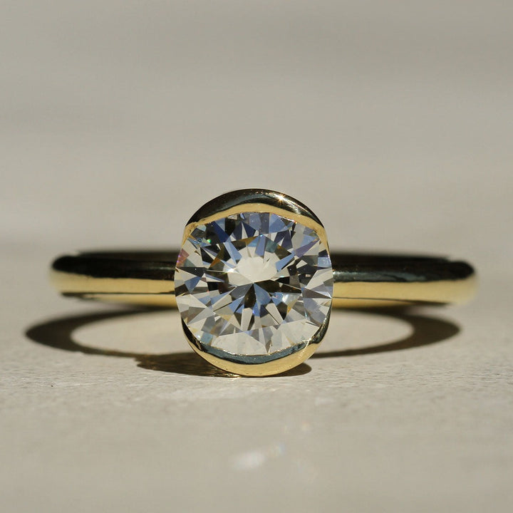 A close-up image of a diamond solitaire ring with a gold band, showcasing the round brilliant cut diamond set in a bezel setting.