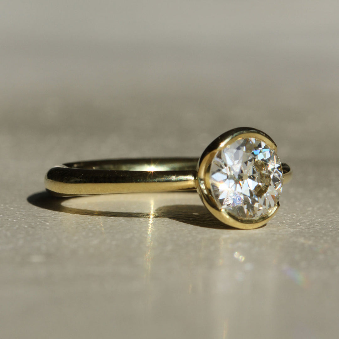 An antique Old European cut solitaire diamond ring with 18k Yellow Gold partial bezel with a tapered band on a neutral background at a slight angle
