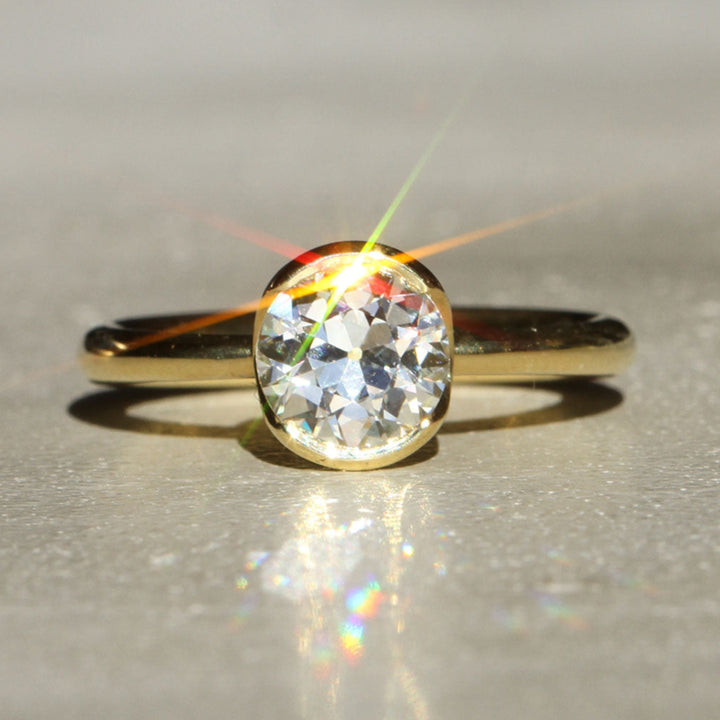 An antique Old European cut solitaire diamond ring with 18k Yellow Gold partial bezel with a tapered band on a neutral background