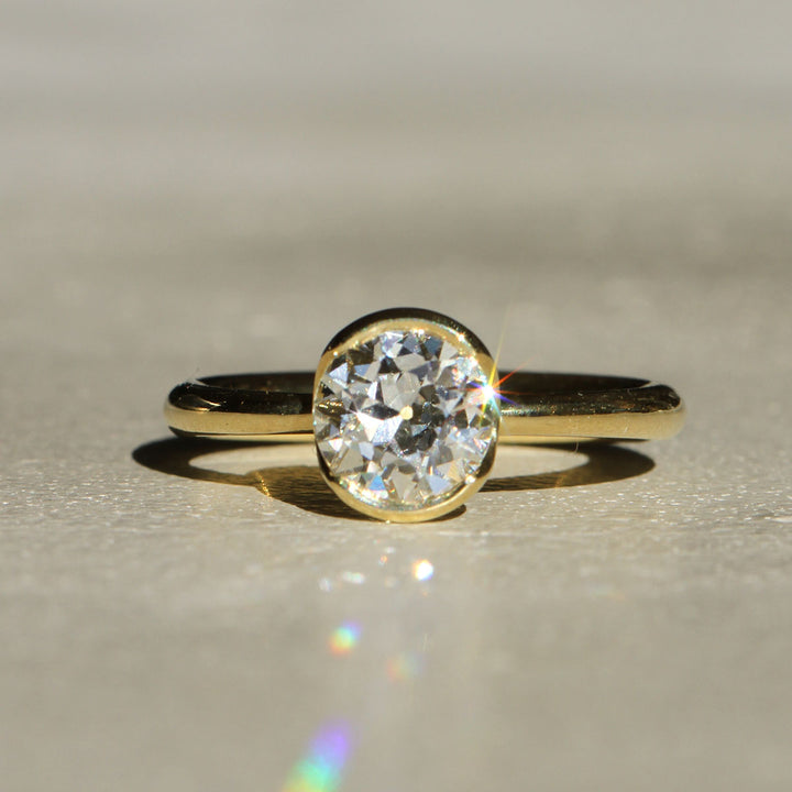 An antique Old European cut solitaire diamond ring with 18k Yellow Gold partial bezel with a tapered band on a neutral background