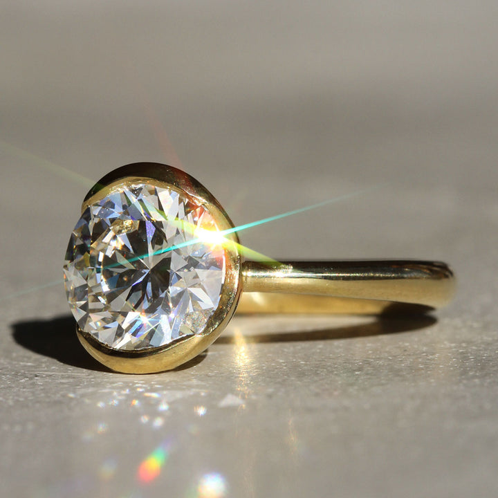 A Round cut diamond solitaire ring with a 18k Yellow Gold partial bezel with a tapered band on a neutral background from a slight angle.