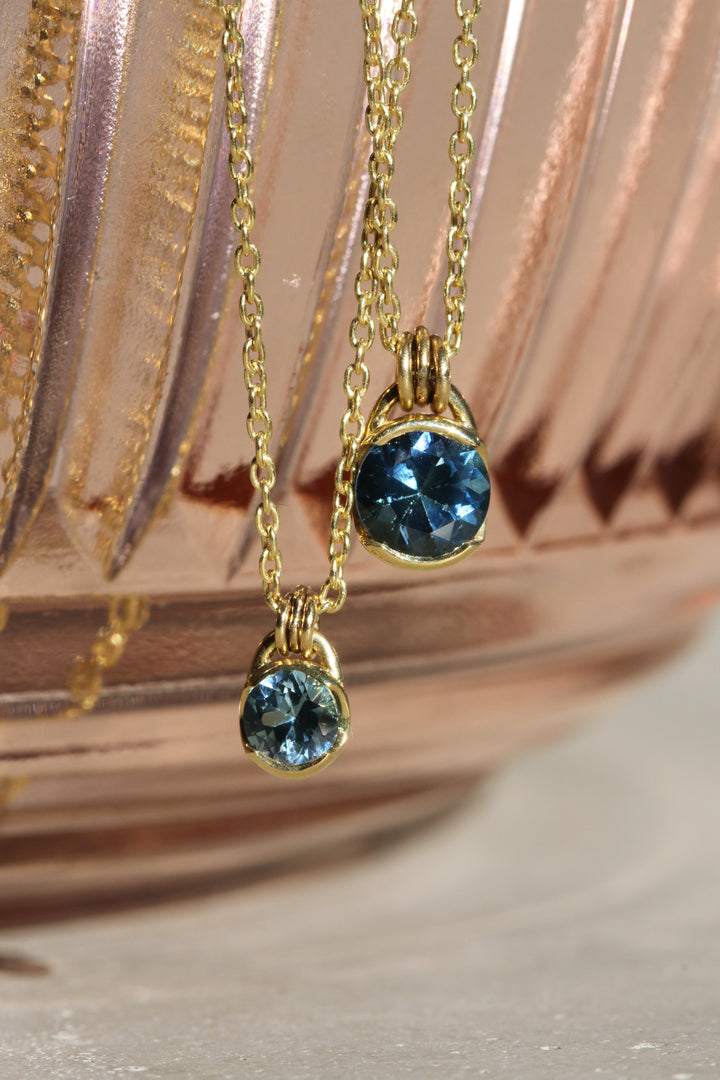 Two dark teal round Montana Sapphire ethical gemstones set in 18k Yellow Gold partial bezel pendants on cable chains connected with three jump rings, hanging from a peach colored glass bowl.