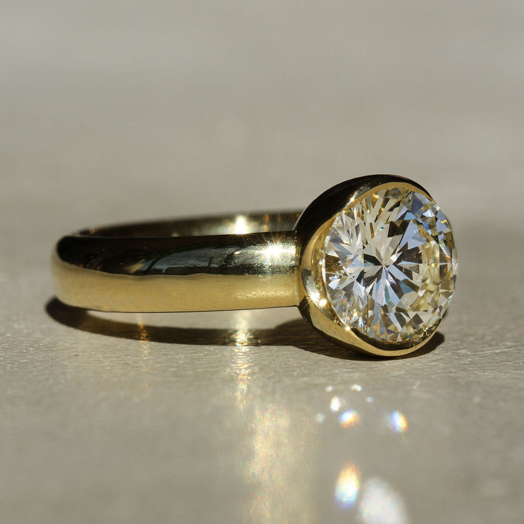 A round brilliant cut white diamond set in a 18k yellow gold partial bezel solitaire ring, with a wide band on a neutral background at a slight angle