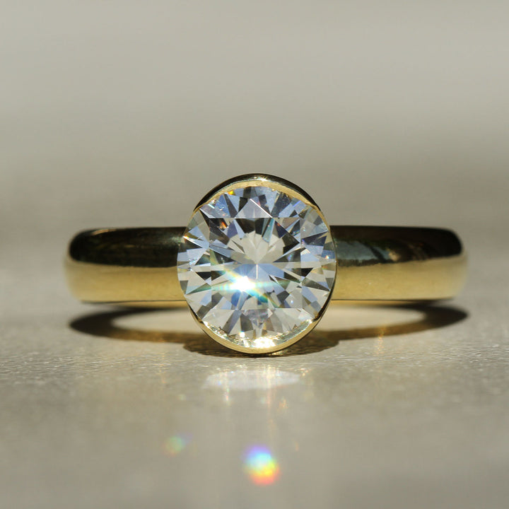 A round brilliant cut white diamond set in a 18k yellow gold partial bezel solitaire ring, with a wide band on a neutral background