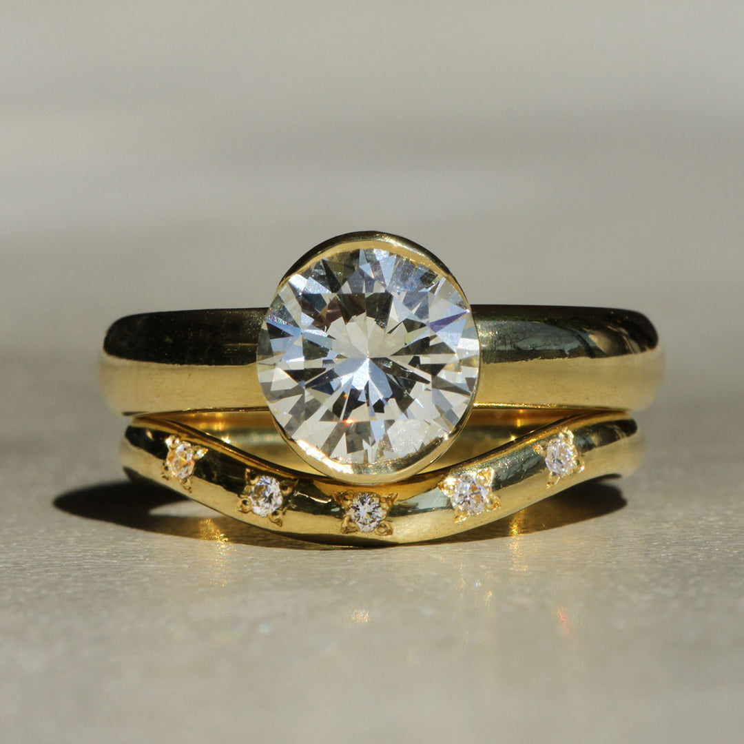 A round brilliant cut white diamond set in a 18k yellow gold partial bezel solitaire ring, with a wide band on a neutral background with a curved diamond band stacked below
