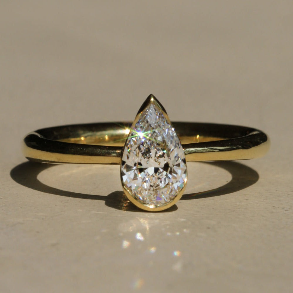 A gold-colored, 18k gold tapered full-round band bezel setting a clear, uncut pear diamond of 0.85 carats.