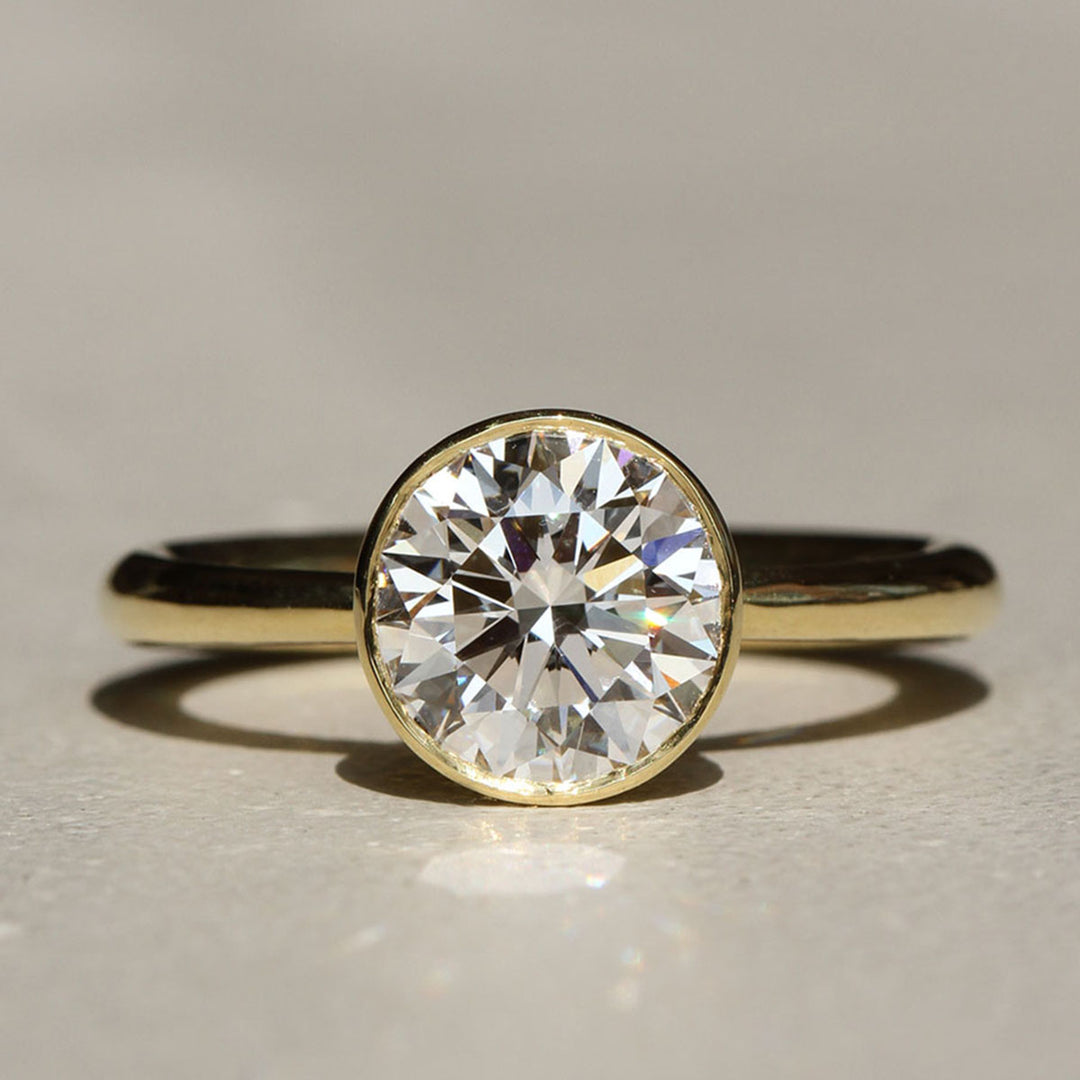 An 18k yellow gold diamond solitaire engagement ring with a round brilliant diamond centerpiece, set in a tulip bezel design, with gentle curves and a polished finish.