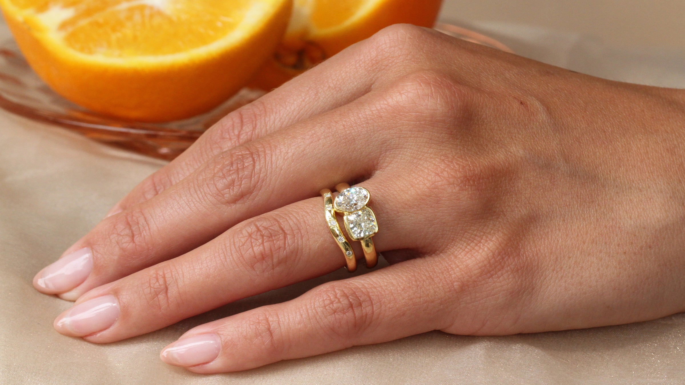 Hand wearing the Sophie Hughes Poppy Bezel Toi et Moi (Cushion & Oval) in 18K Yellow Gold, styled with the SH Shallow Curved Sprinkle Band in 18K Yellow Gold, with oranges in the background
