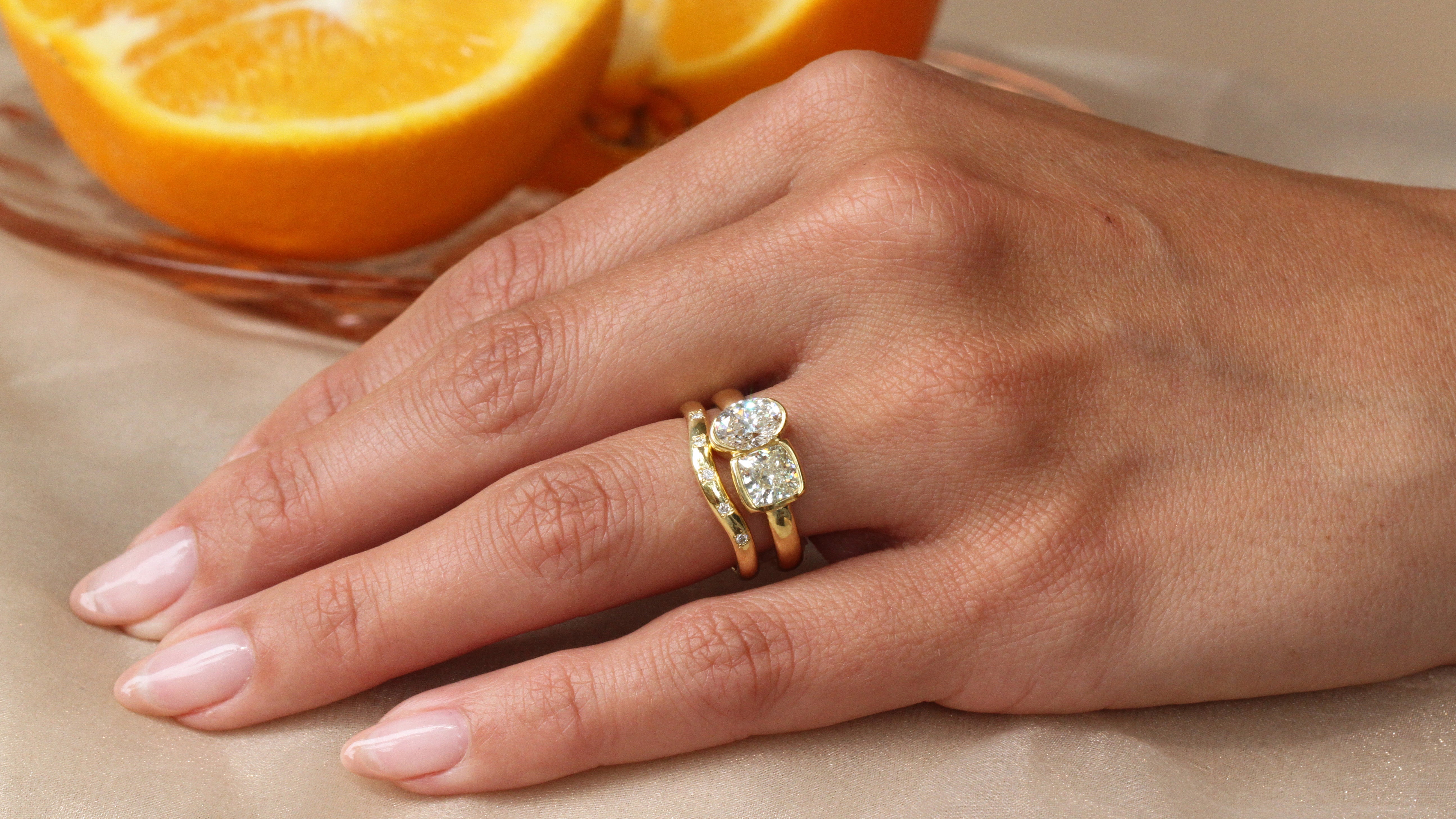 Hand wearing the Sophie Hughes Poppy Bezel Toi et Moi (Cushion & Oval) in 18K Yellow Gold, styled with the SH Shallow Curved Sprinkle Band in 18K Yellow Gold, with oranges in the background