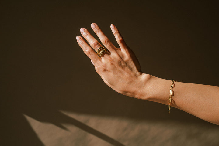 A person's hand displaying a thin link chain bracelet with a subtle hammered texture, wearing a ring, against a dark background.