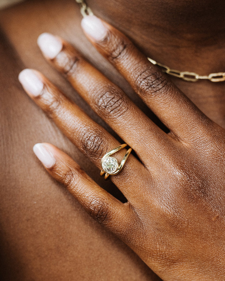 A hand wearing An 18k yellow gold solitaire diamond ring featuring an antique Old European cut diamond with a unique looped double band design.