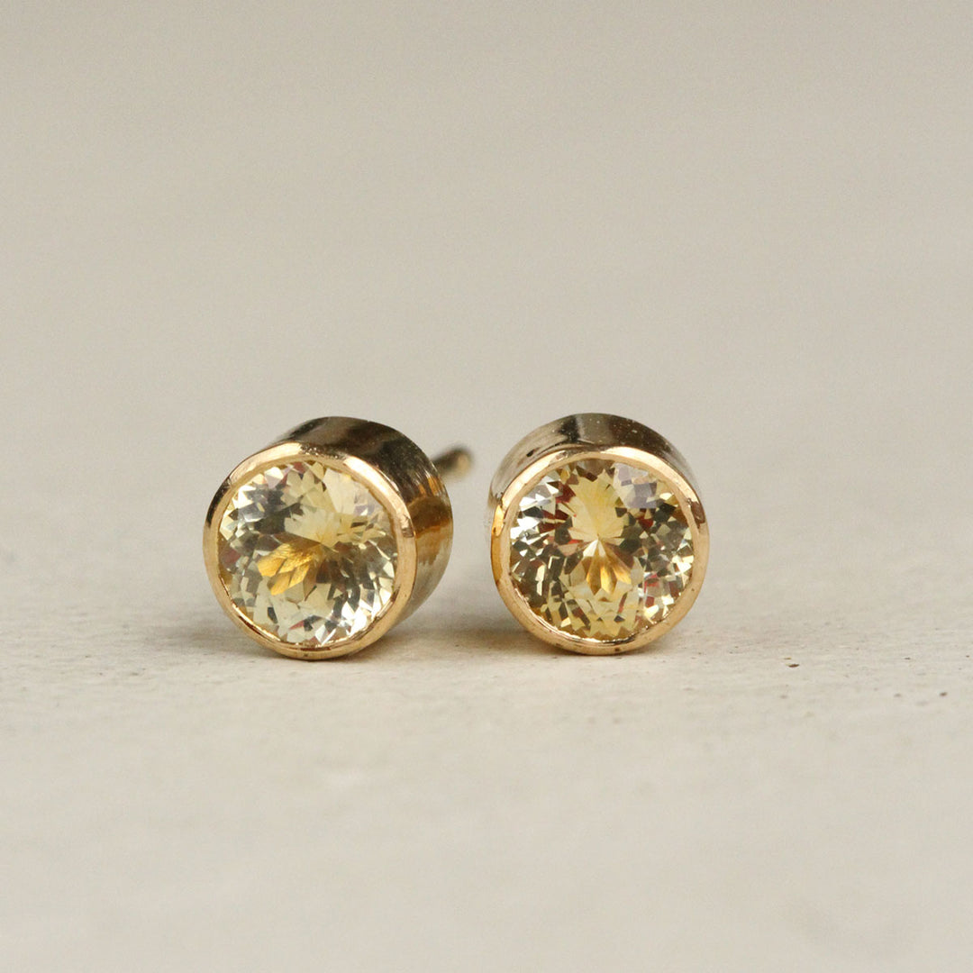 A pair of yellow gold bezel stud earrings featuring yellow Montana Sapphires, set against a beige background.